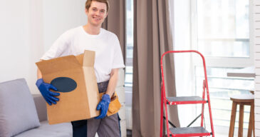 Smiling male worker of moving and delivery company holding cardboard box. Loader in overalls posing against background of colleague who packs cardboard boxes. Moving service concept. Smiling male worker of moving and delivery company holding cardboard box. Loader in overalls posing against background of colleague who packs cardboard boxes. Moving service concept